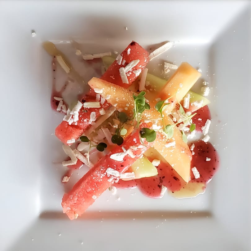 Colorful fruit salad with watermelon, cantaloupe, honeydew, microgreens, and cheese crumbles on a white plate.