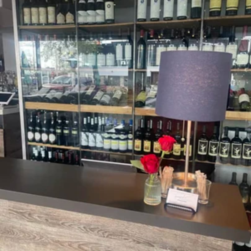 A dark wooden counter with a lamp and red roses, backed by shelves of wine bottles.
