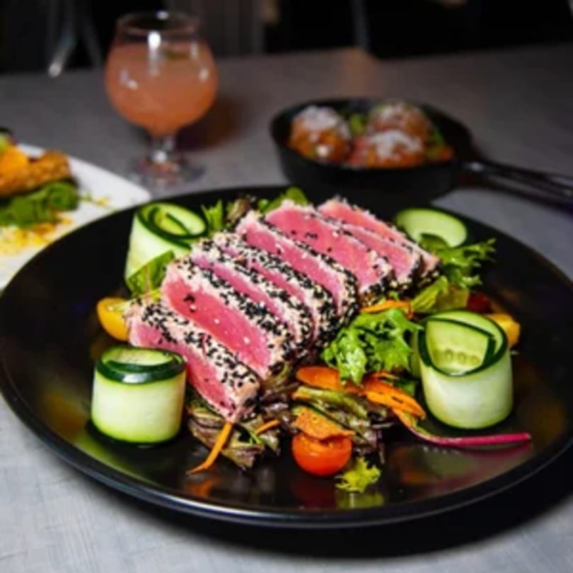Seared tuna with sesame seeds, mixed greens, cherry tomatoes, and cucumber on a black plate.