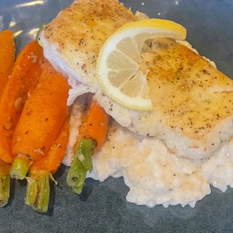 Pan-seared chicken with a lemon slice, creamy risotto, and roasted carrots on a dark plate.
