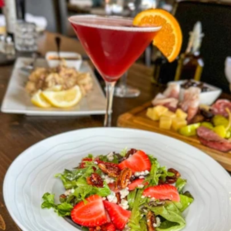 A vibrant red cocktail, strawberry salad, calamari, and charcuterie board on a wooden table.