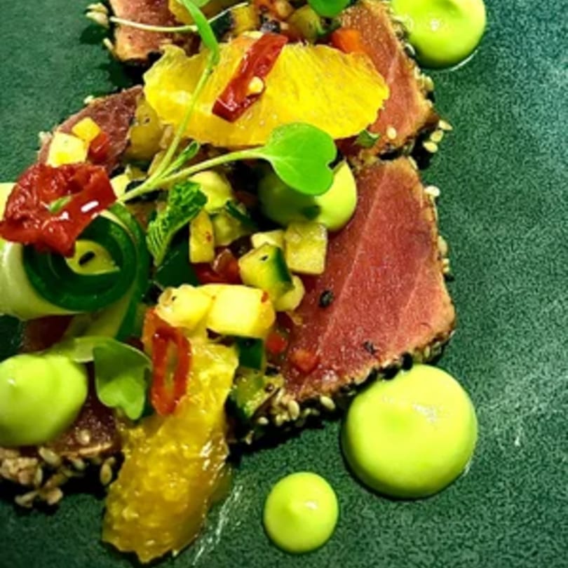 Seared tuna with sesame seeds, citrus, diced vegetables, and green sauce on a dark plate.